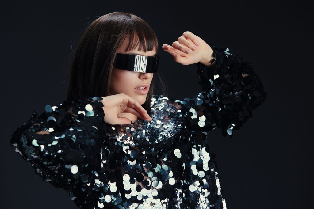 A stylish woman in sunglasses and a sequinned shirt stands confidently with hands on her hips and