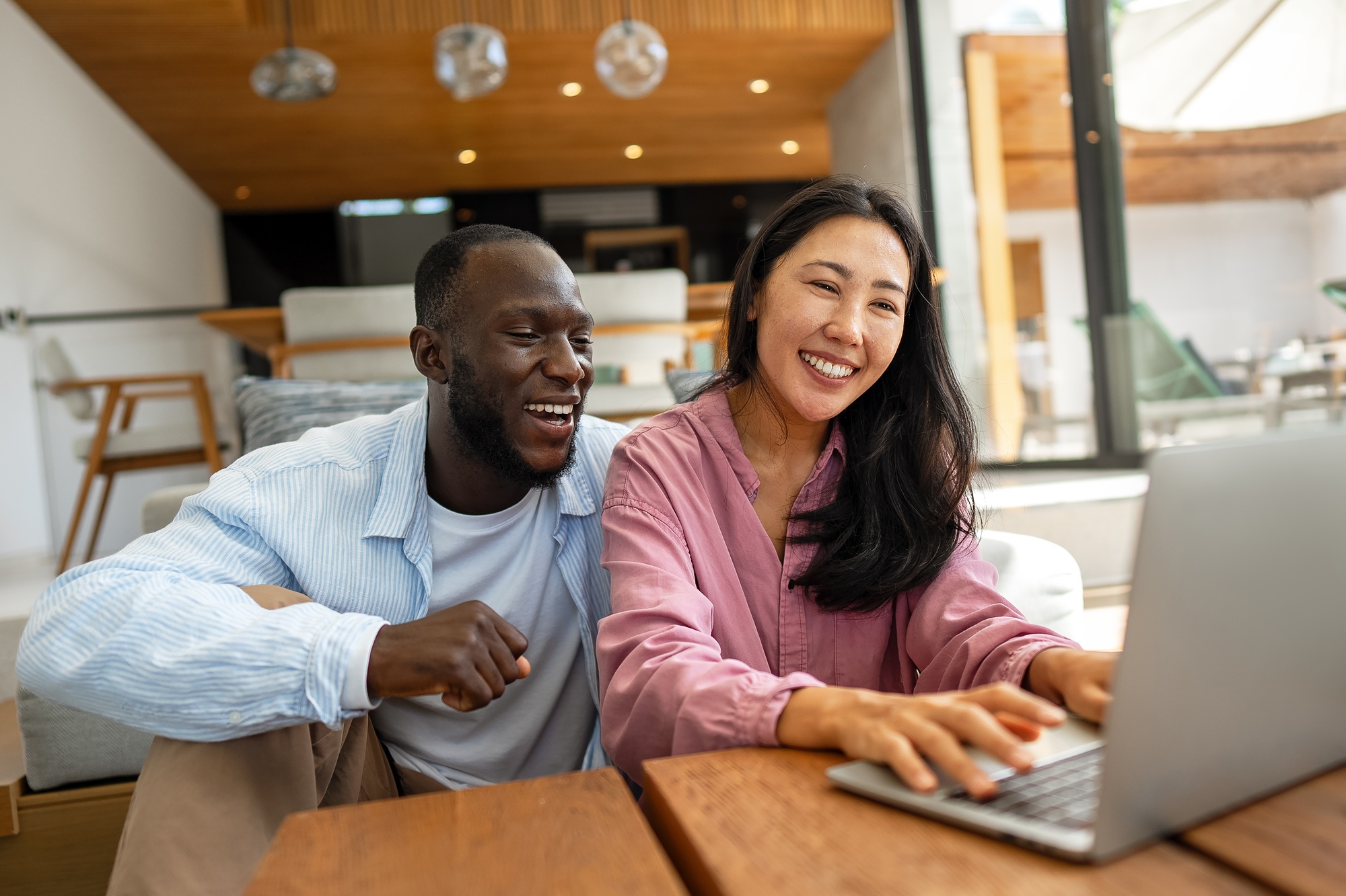 Happy young diverse, multiethnic couple planning budget, reading good news on laptop at home
