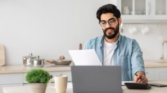 Portrait of happy arab guy reading insurance documents at home, sitting at table in kitchen and