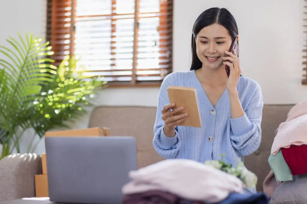 Portrait young attractive hipster asian female owner startup call phone with box at home prepare