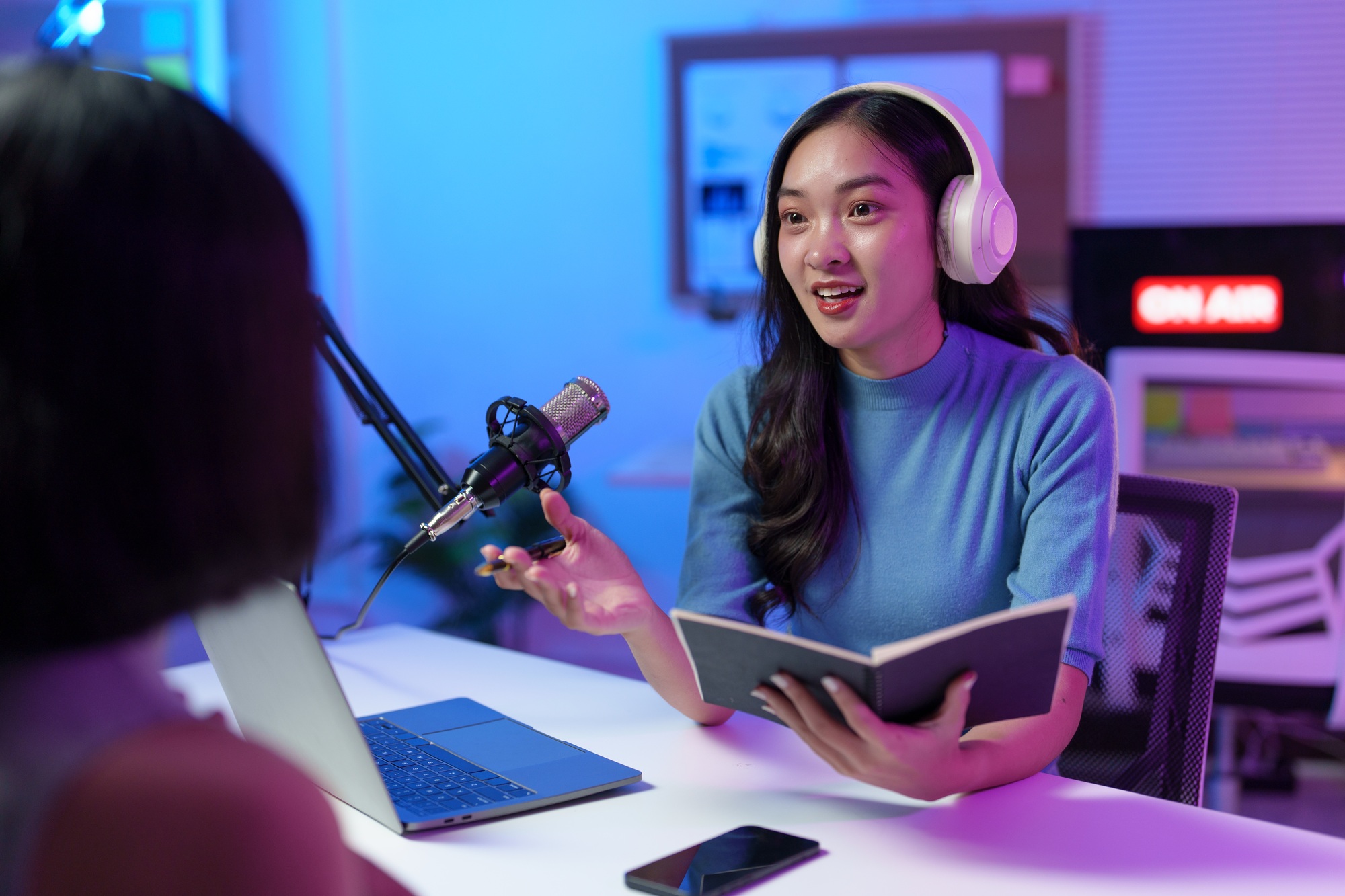 Two female podcasters are recording a show in a professional studio, using a laptop, microphone, and